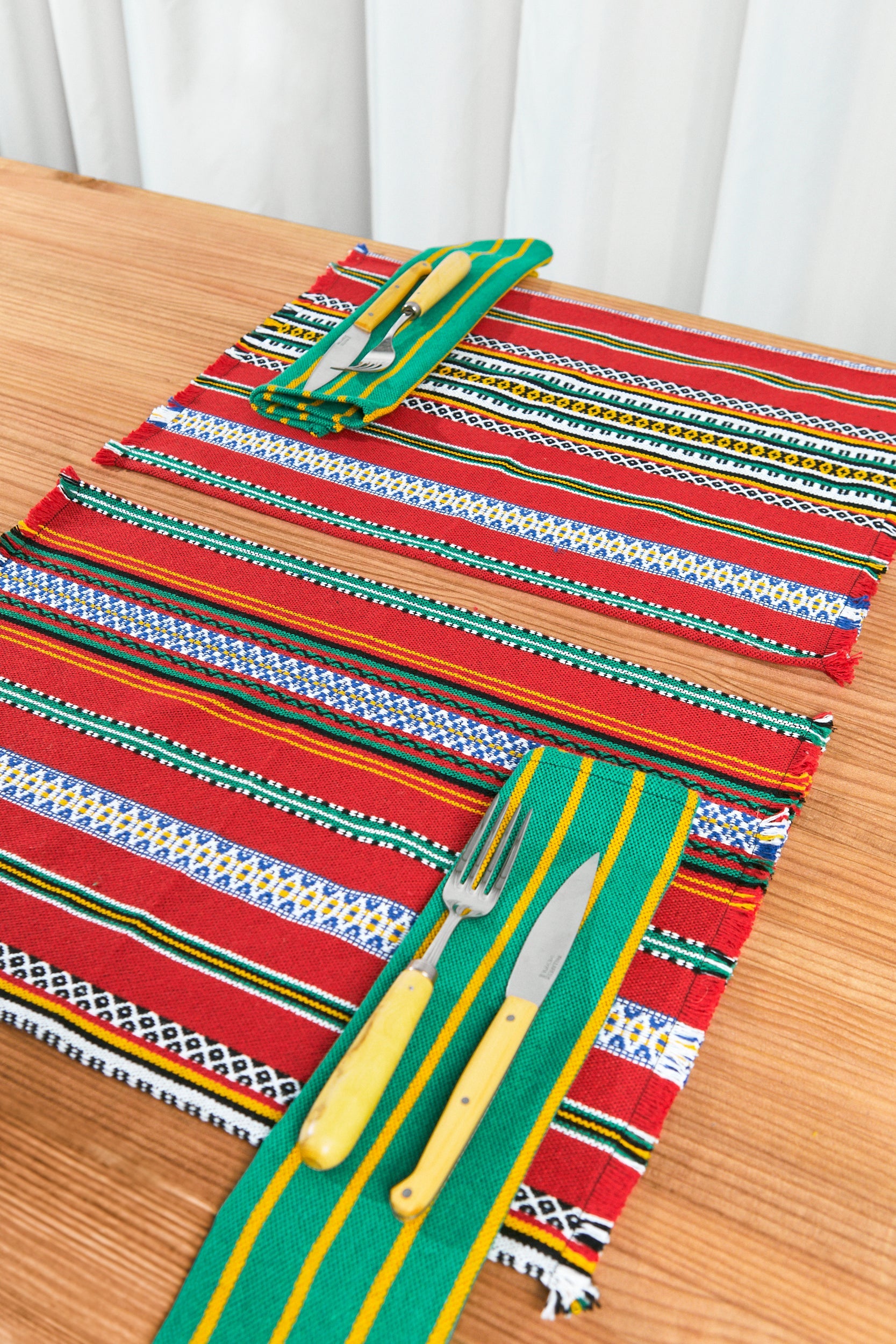 Mantel tejido en tonos rojo, verde y amarillo, con cubiertos de acero y mangos amarillos, estilo artesanal.