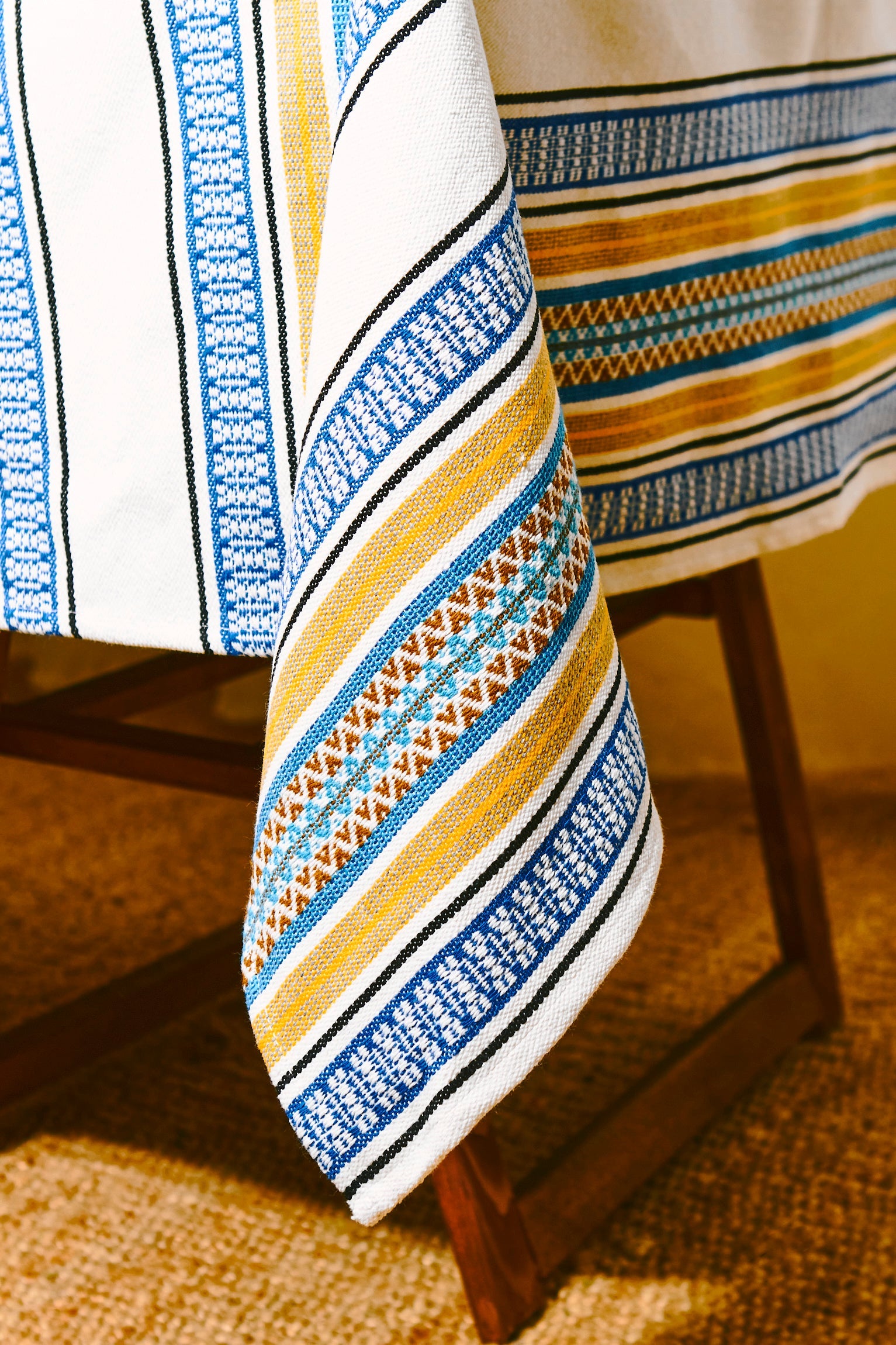 Mantel de algodón blanco con estampado azul, amarillo y marrón; estilo artesanal y elegante para decoración.