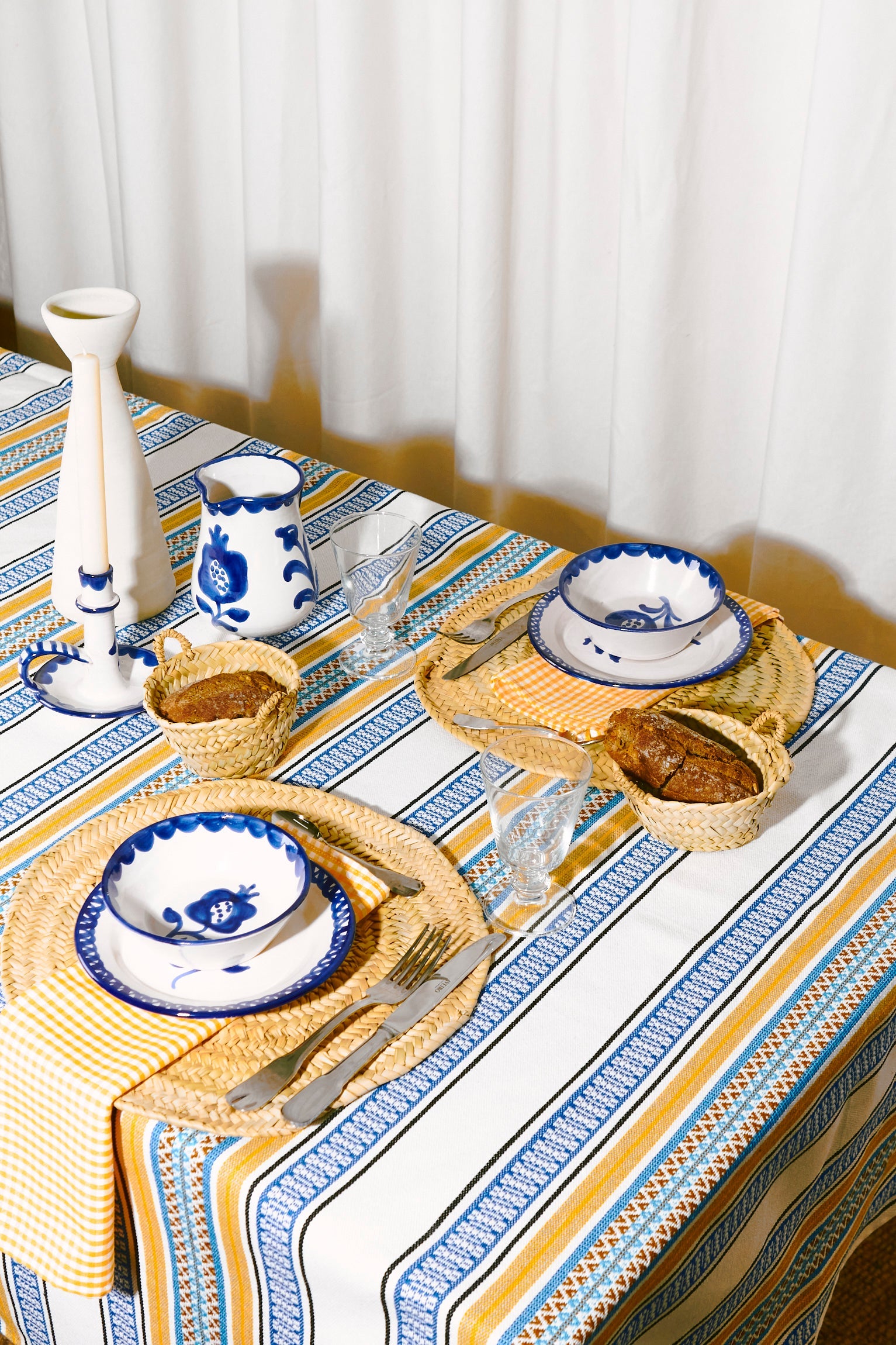 Juego de mesa con mantelería azul y amarilla, cerámica blanca decorada, y elementos de mimbre.