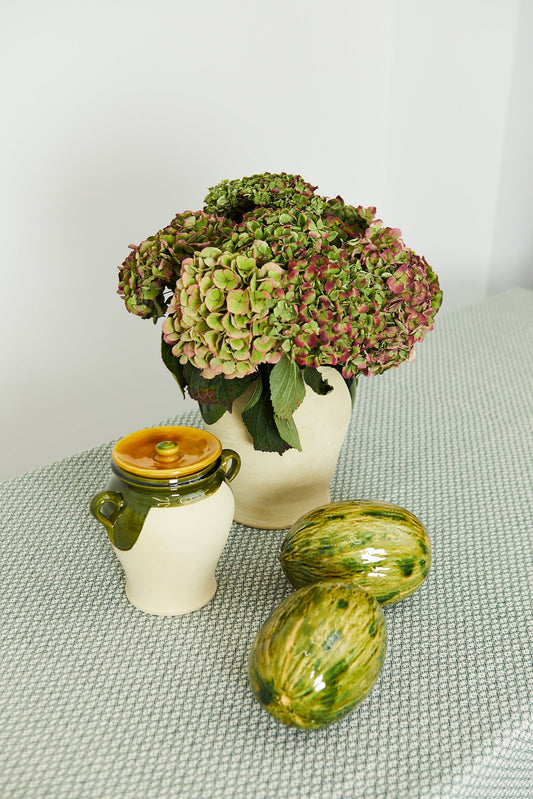 Ramo de hortensias verdes y moradas en jarrón beige, acompañado de tarro amarillo y frutas decorativas verdes.