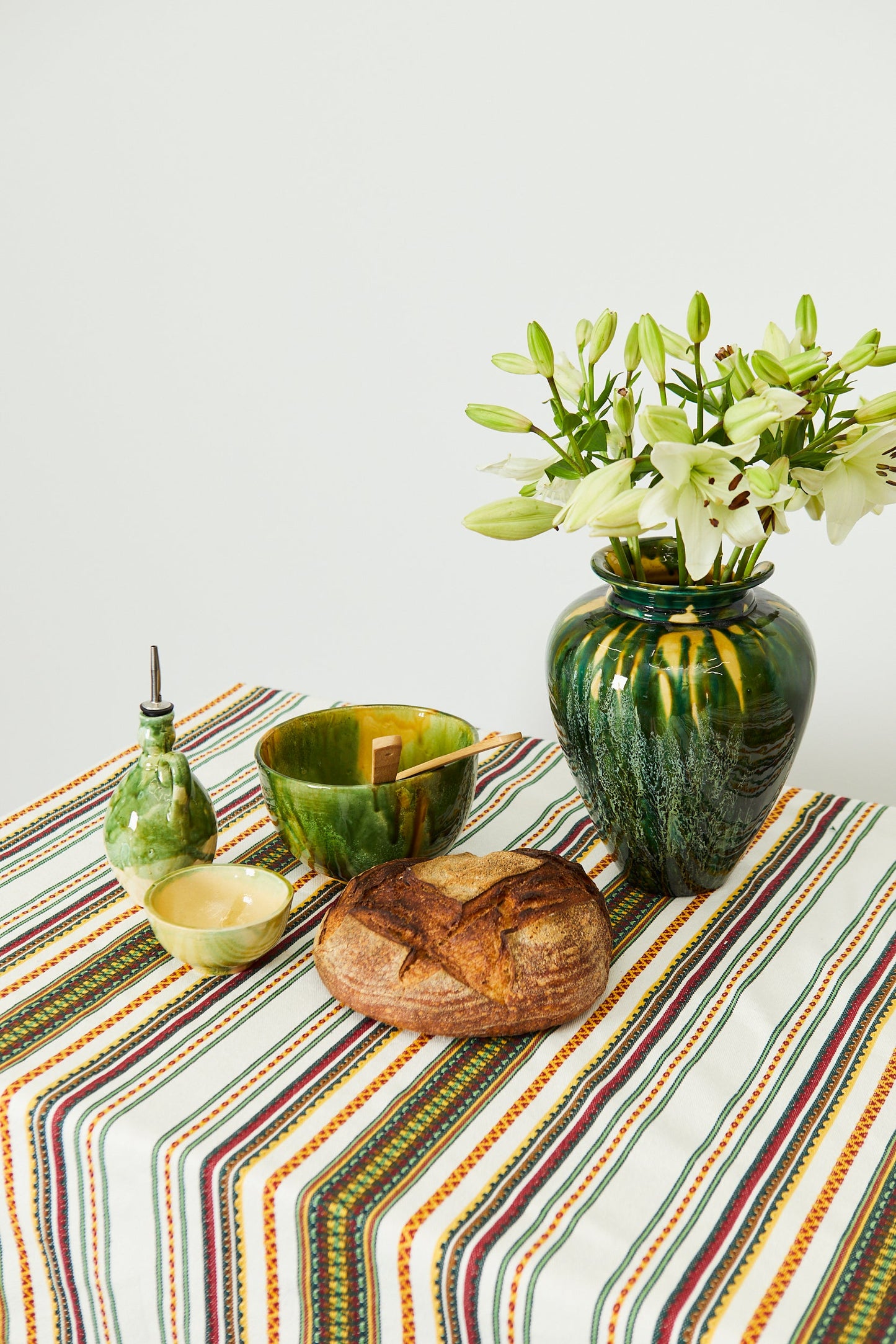 Jarrón verde con flores, cuencos de cerámica amarillos y verdes, pan artesanal sobre mantel rayado multicolor.
