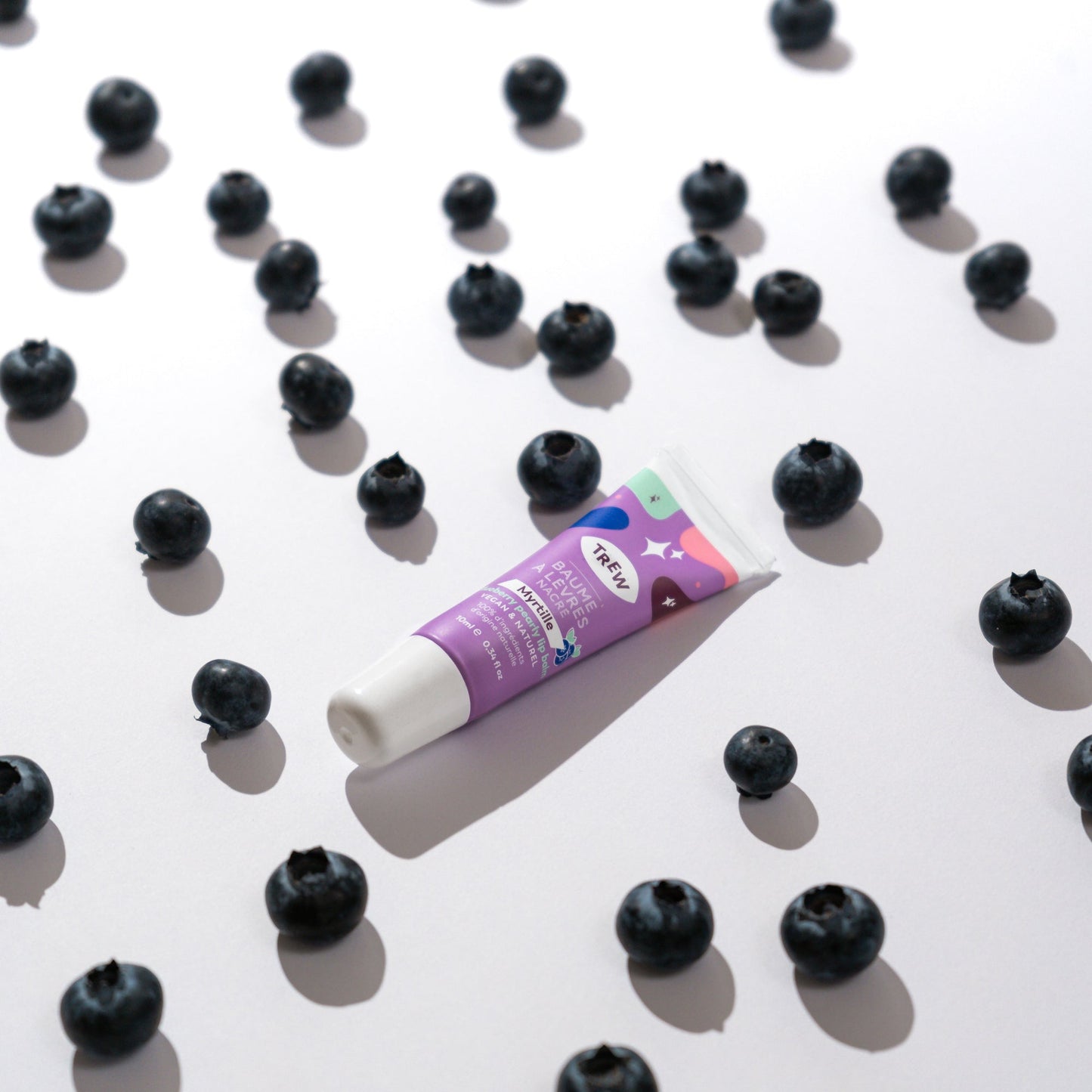A tube of purple blueberry tinted lip balm centered among scattered fresh blueberries on a white background.