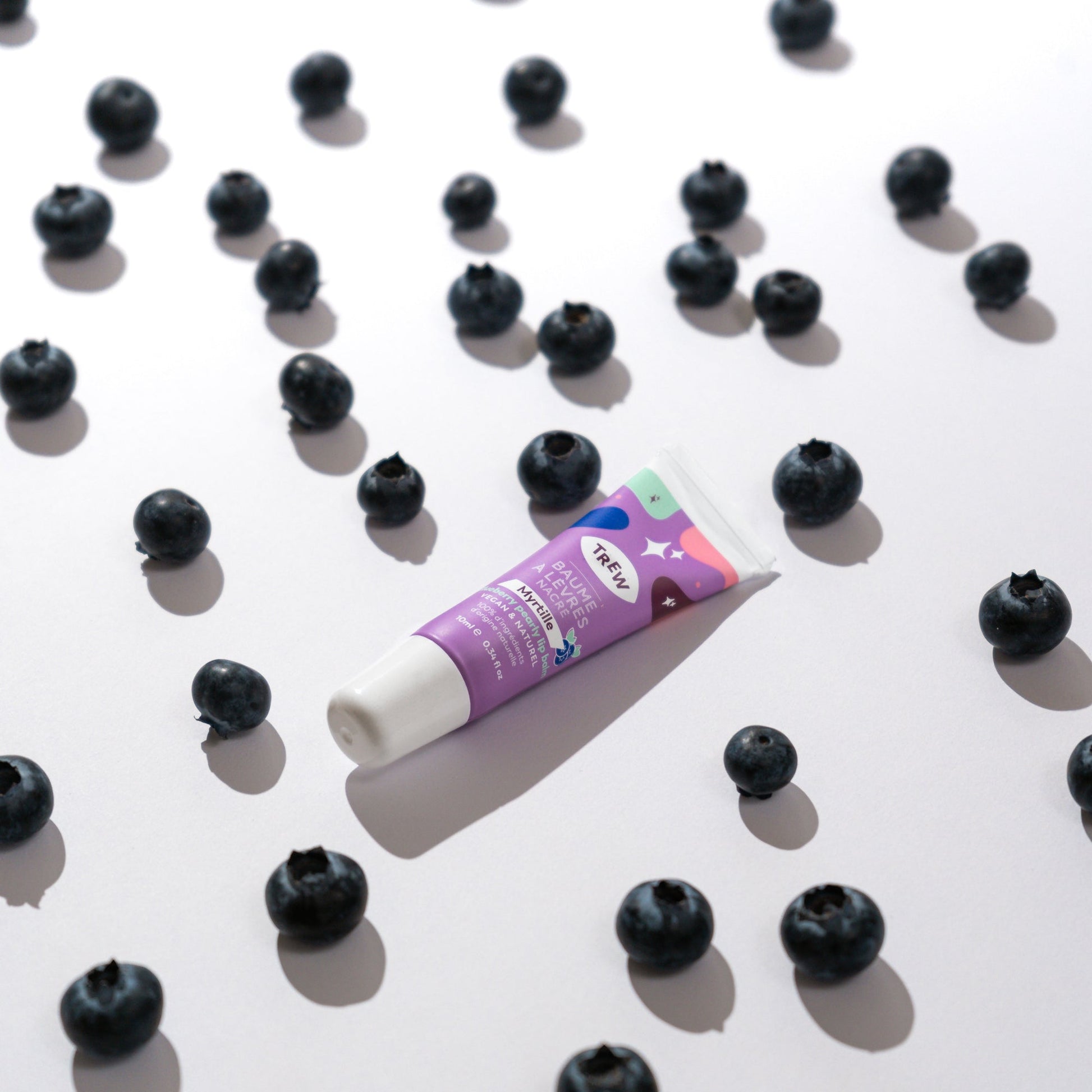 A tube of purple blueberry tinted lip balm centered among scattered fresh blueberries on a white background.