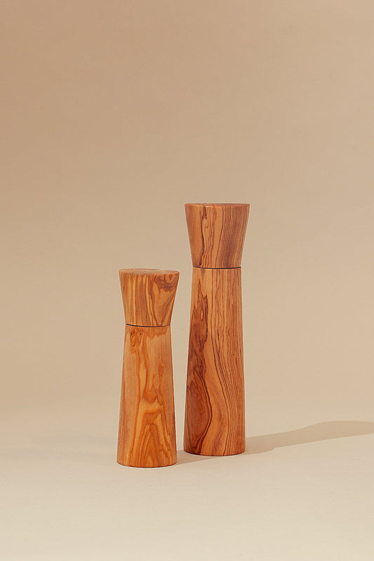 Dos molinillos de madera de olivo con veteado natural y diseño minimalista sobre fondo color arena.