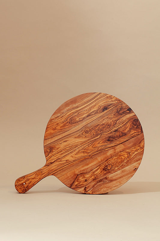 Tabla redonda de madera de olivo con mango, mostrando sus vetas naturales y tonos marrones sobre un fondo beige.