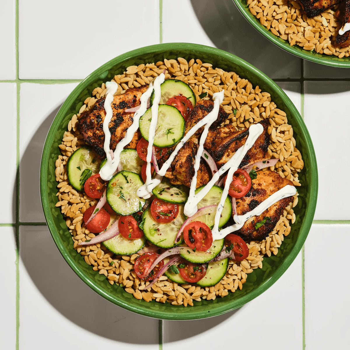 A green bowl of Mediterranean rice, grilled chicken, sliced cucumbers, tomatoes, and red onion, drizzled with white sauce.