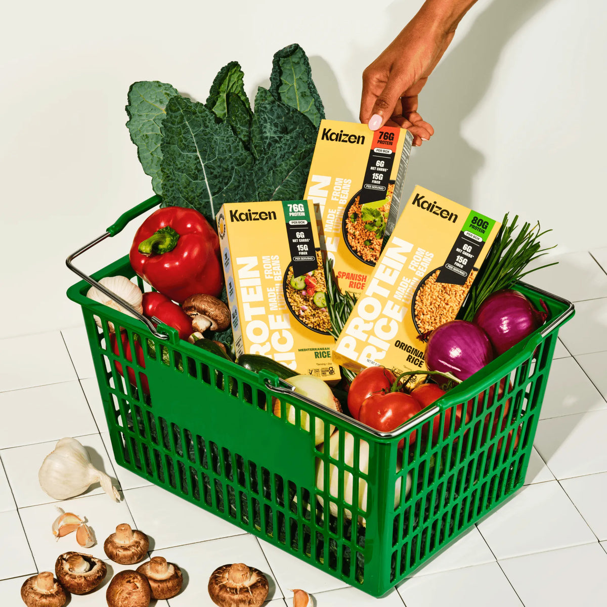 Green shopping basket filled with boxes of Kaizen protein rice, fresh kale, bell peppers, onions, and mushrooms.