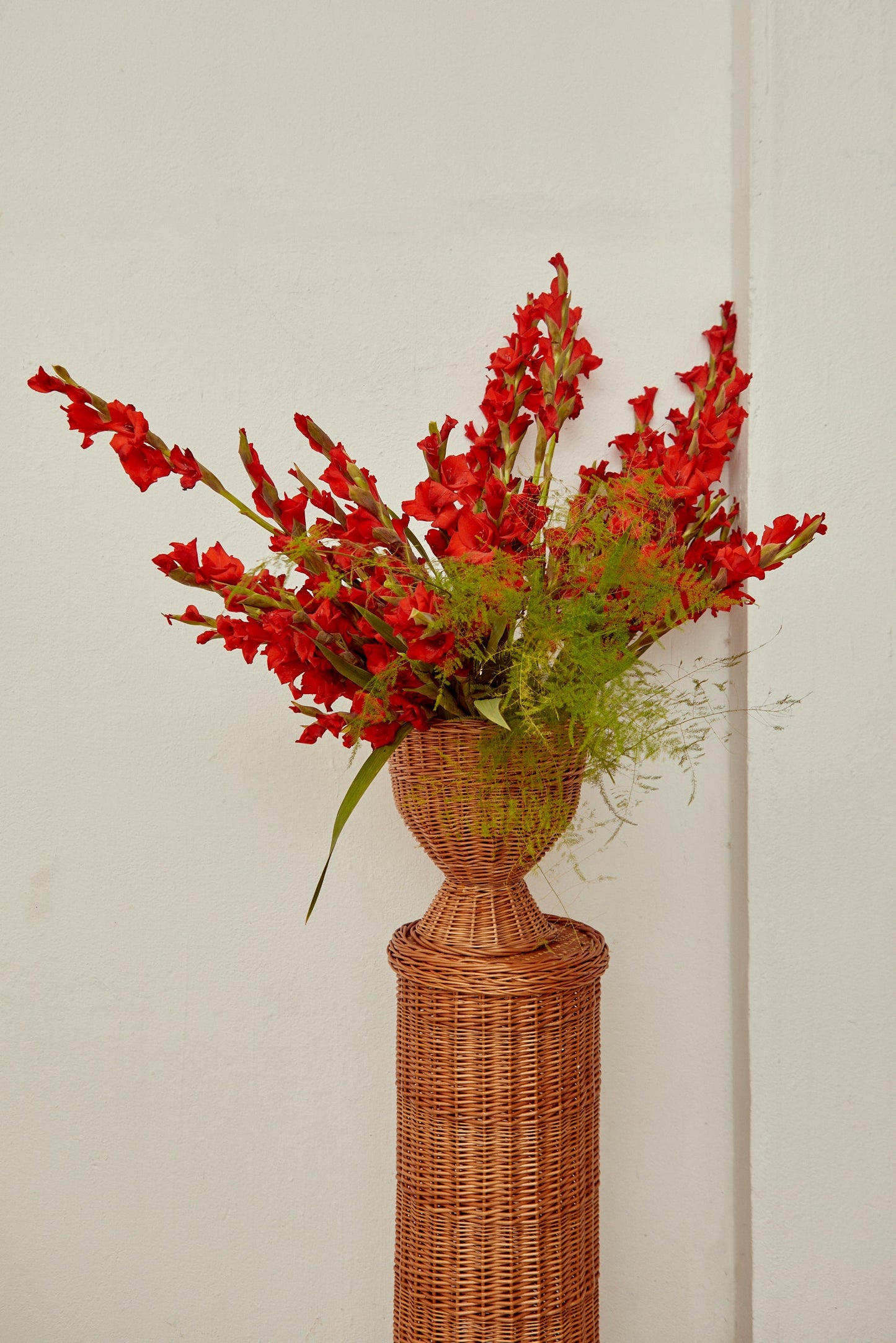 Jarrón de mimbre con flores rojas de gladiolos y hojas verdes, estilo rústico, decorativo para el hogar.