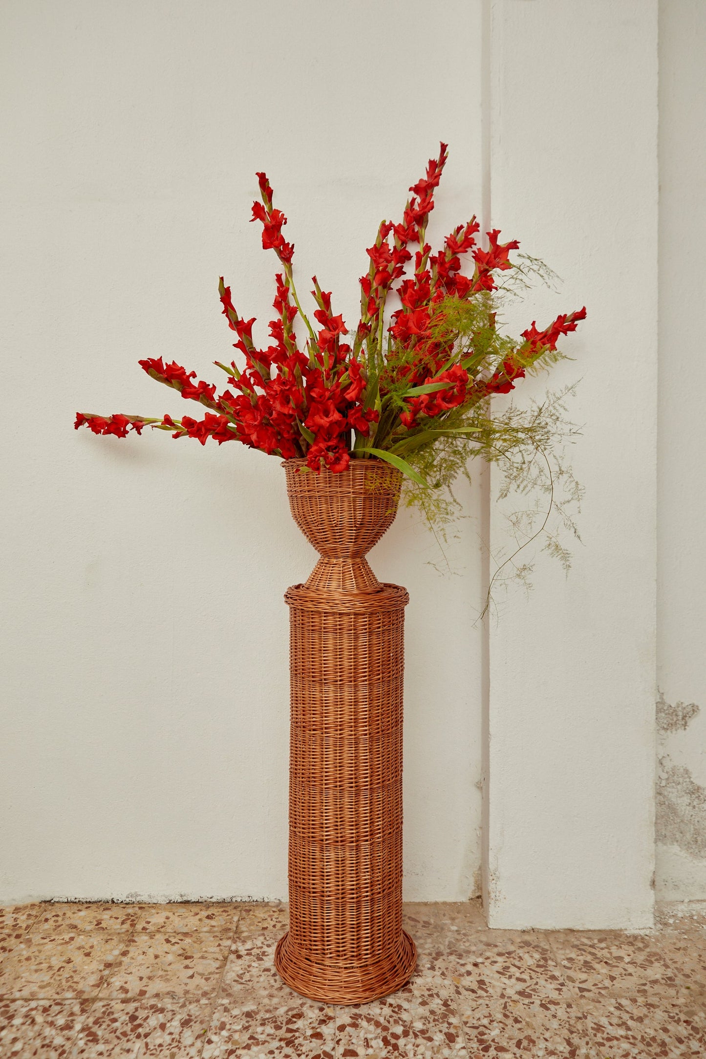 Jarrón de mimbre trenzado en color marrón con flores rojas y verdes, estilo bohemio, ideal para decoración.