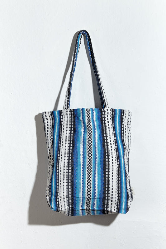 Tote bag de tela con estampado de rayas verticales y patrones geométricos en tonos azules, blancos y negros.