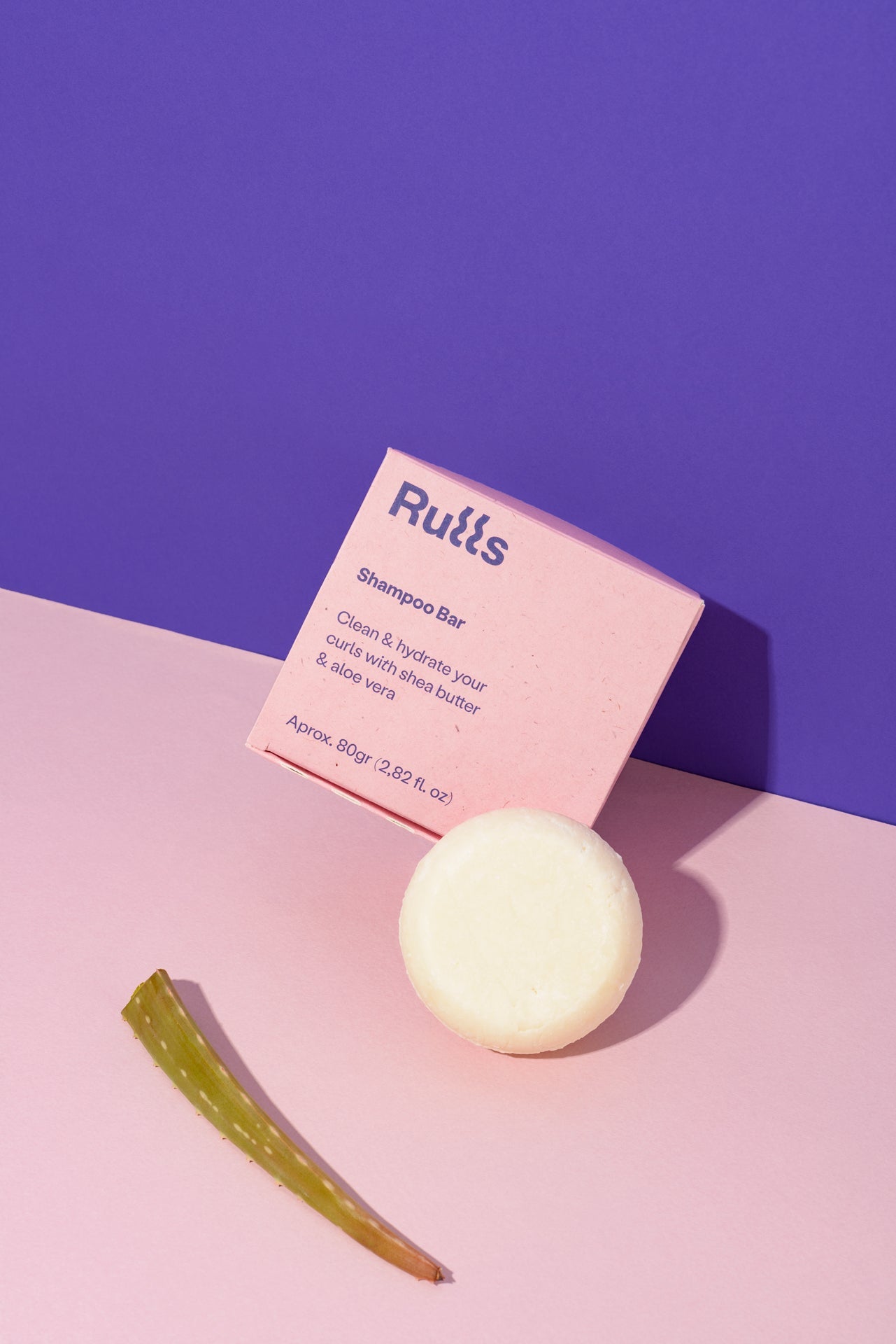Barra de champú blanca con envoltura rosa, contiene manteca de karité y aloe vera, para cabello rizado.