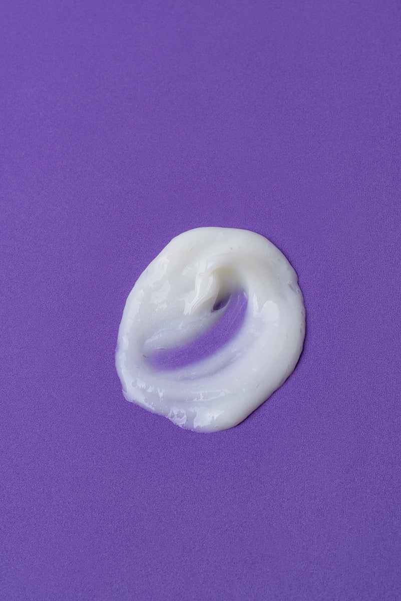 Crema hidratante blanca sobre fondo púrpura, textura suave y cremosa, ideal para el cuidado de la piel.