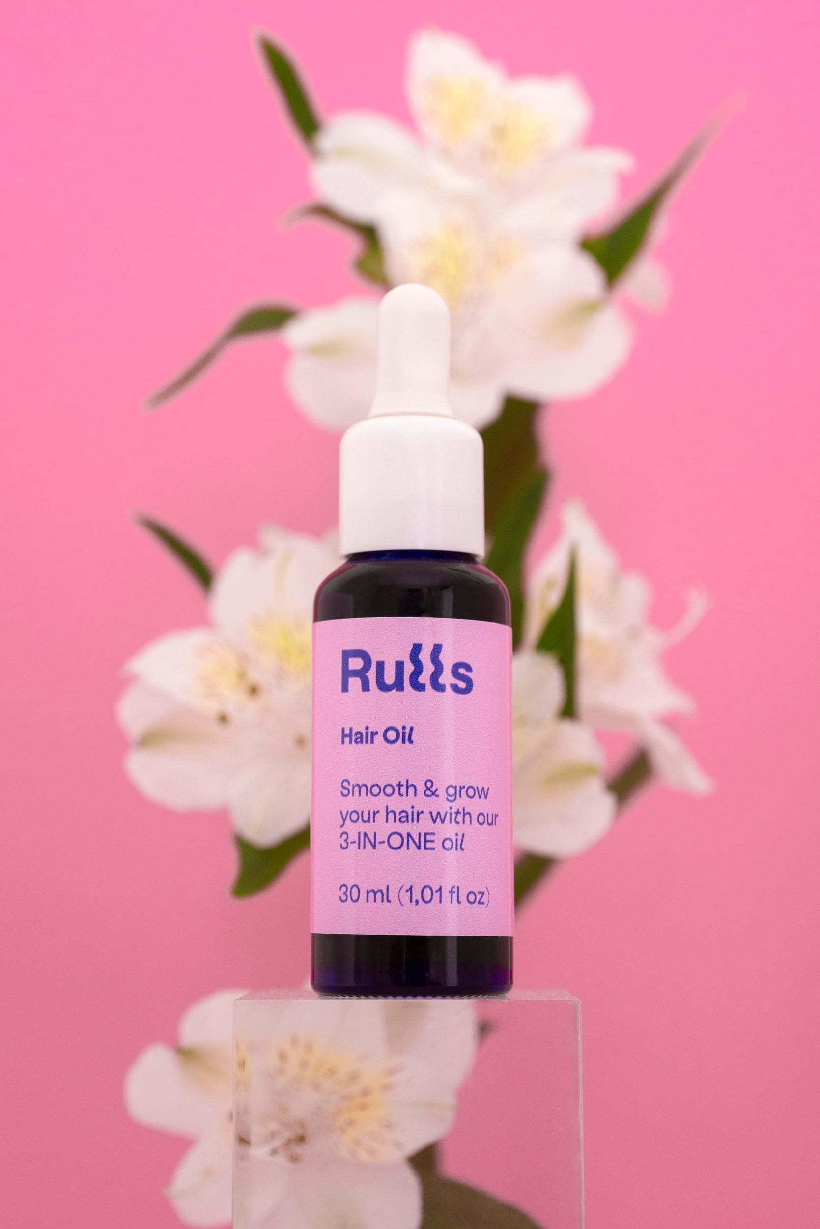 Aceite para el cabello Rulls 30 ml, frasco negro con etiqueta morada, fondo rosa y flores blancas.