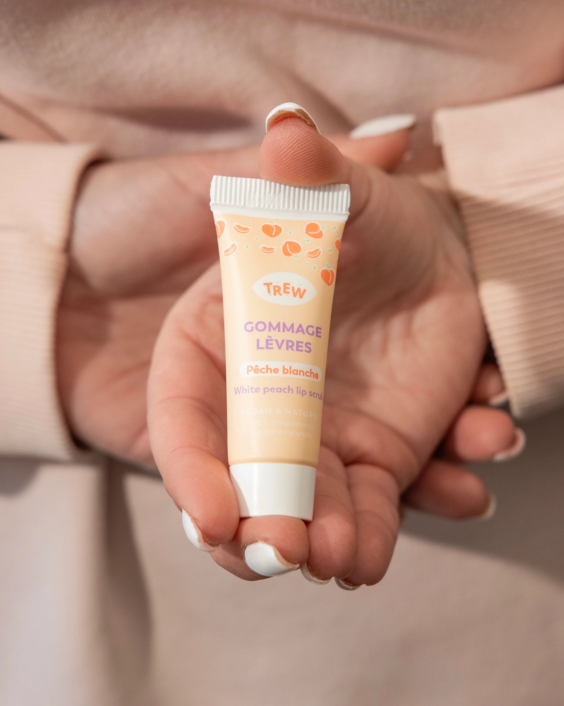 Mains tenant un petit tube de gommage lèvres Trew pêche blanche aux tons orangés, sur fond beige assorti.