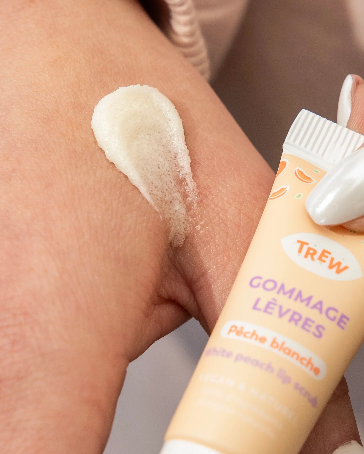 Texture granuleuse de gommage pour les lèvres Trew au parfum pêche, appliquée sur la peau près du tube orangé.