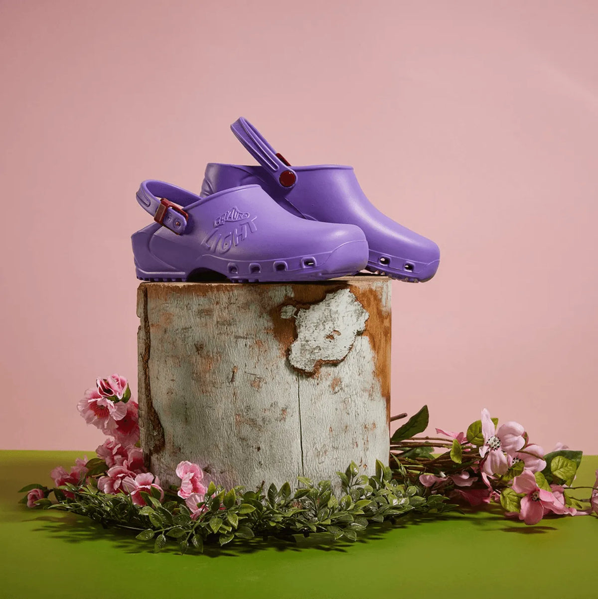 Lilac rubber clogs with heel straps and side ventilation holes, displayed on a wooden stump with flowers.