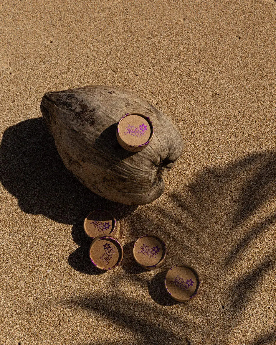 Round, eco-friendly tan cosmetic containers with purple logo branding arranged on beach sand with a whole coconut.