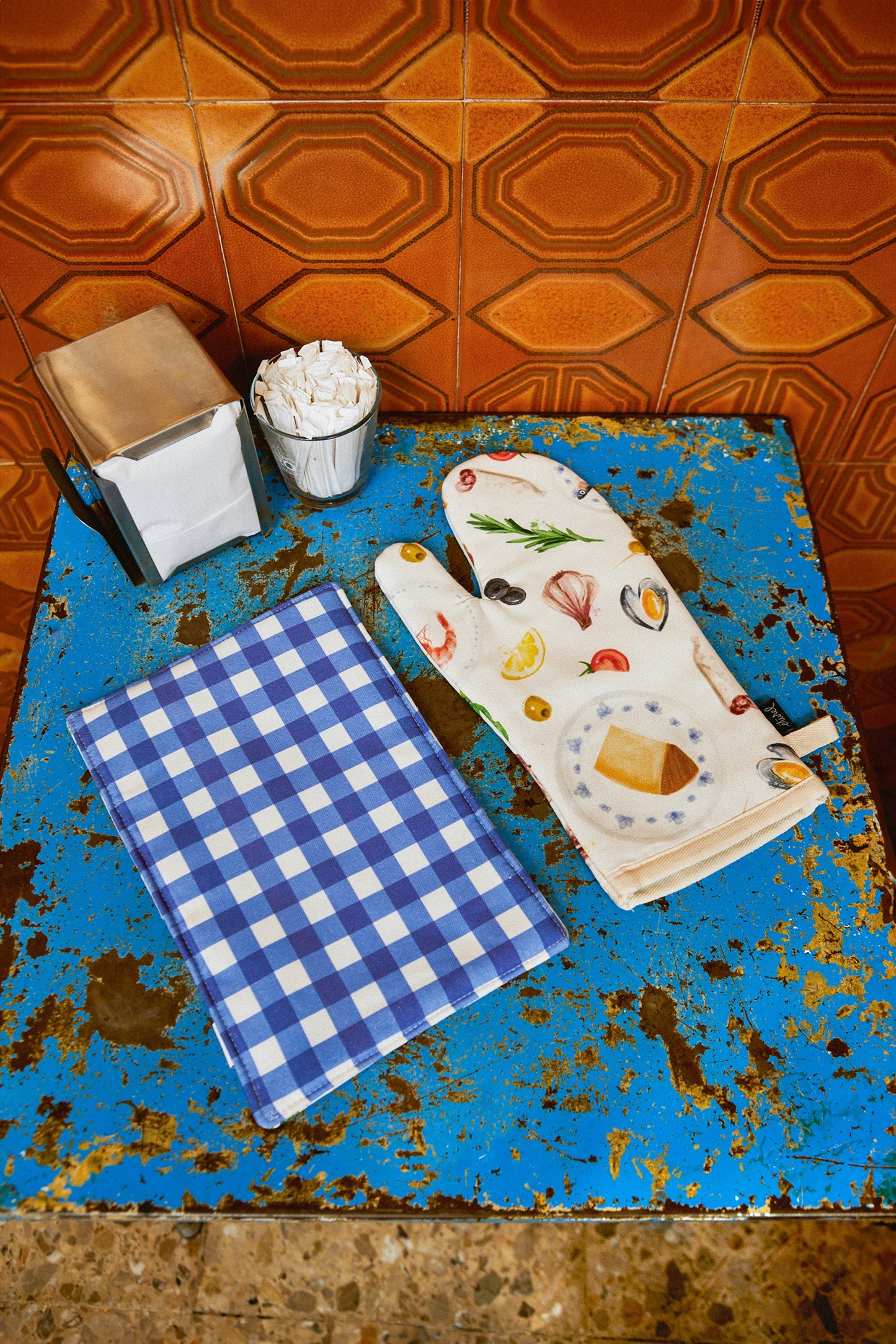 Manopla de cocina blanca con estampado de alimentos y paño de cuadros azules sobre mesa azul.