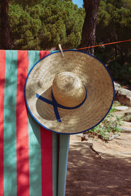 Sombrero de paja con cinta azul, colgado sobre toalla de playa rayada en rojo y verde, estilo veraniego.