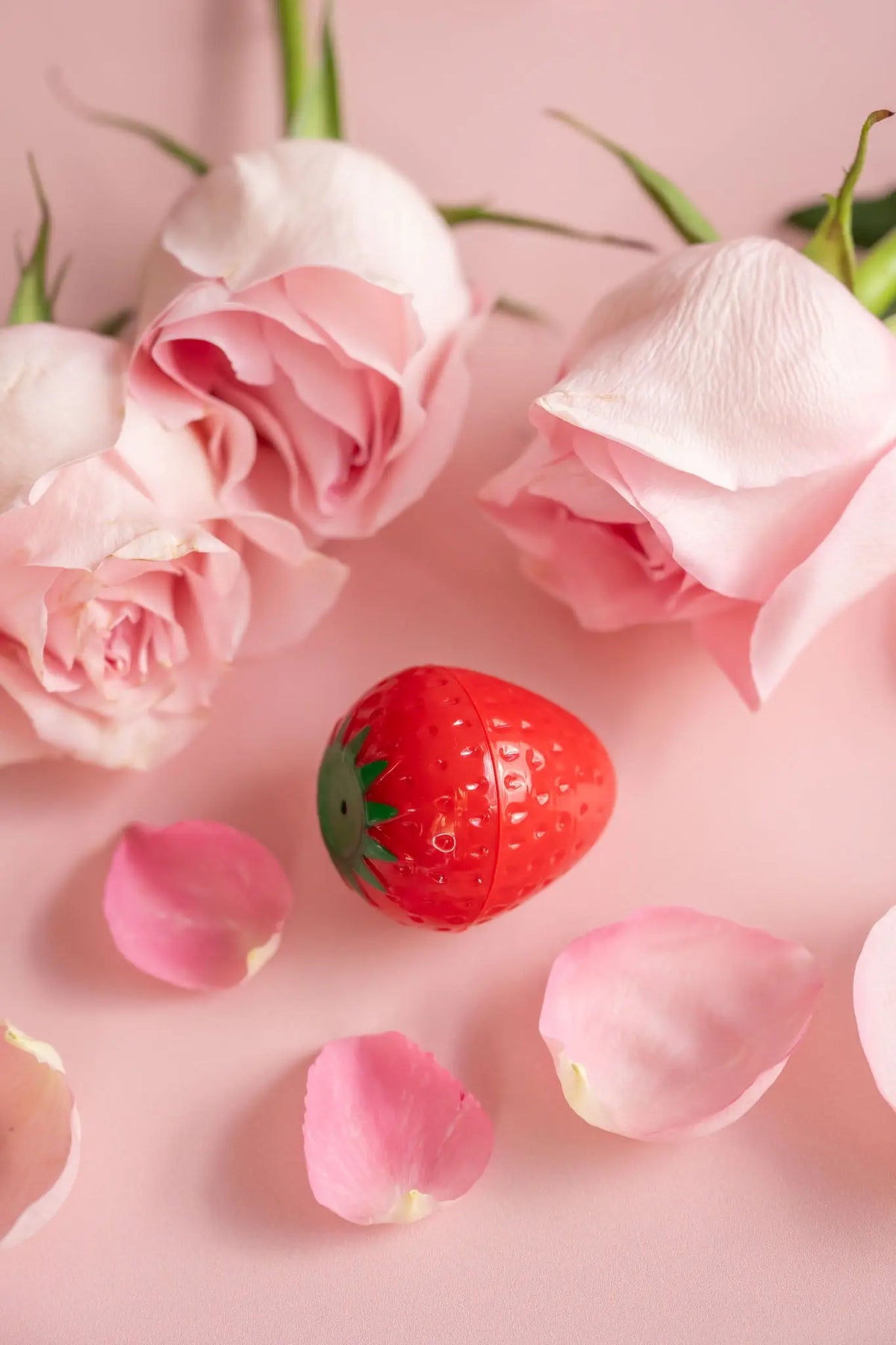 Strawberry-shaped red lip butter container surrounded by soft pink roses and scattered petals on a pink background.