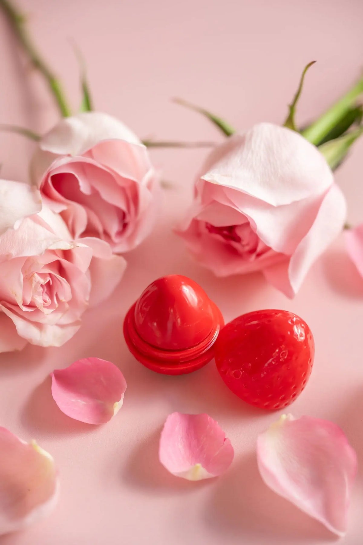 Strawberry-shaped red lip butter with its cap off, surrounded by light pink rose buds and petals on a pink background.