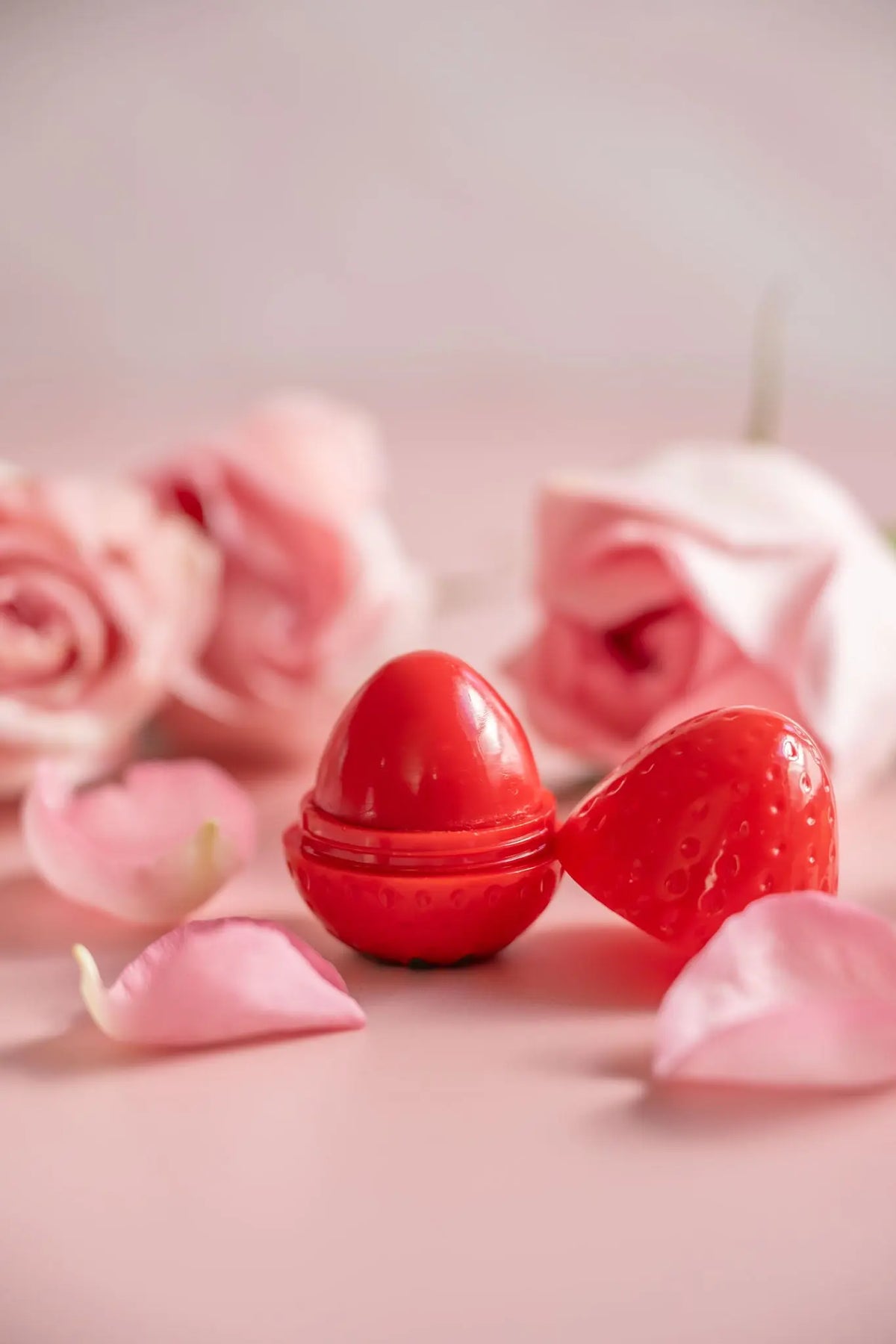 Strawberry-shaped red lip butter container open on a pink background with soft-focus pale pink rose petals.