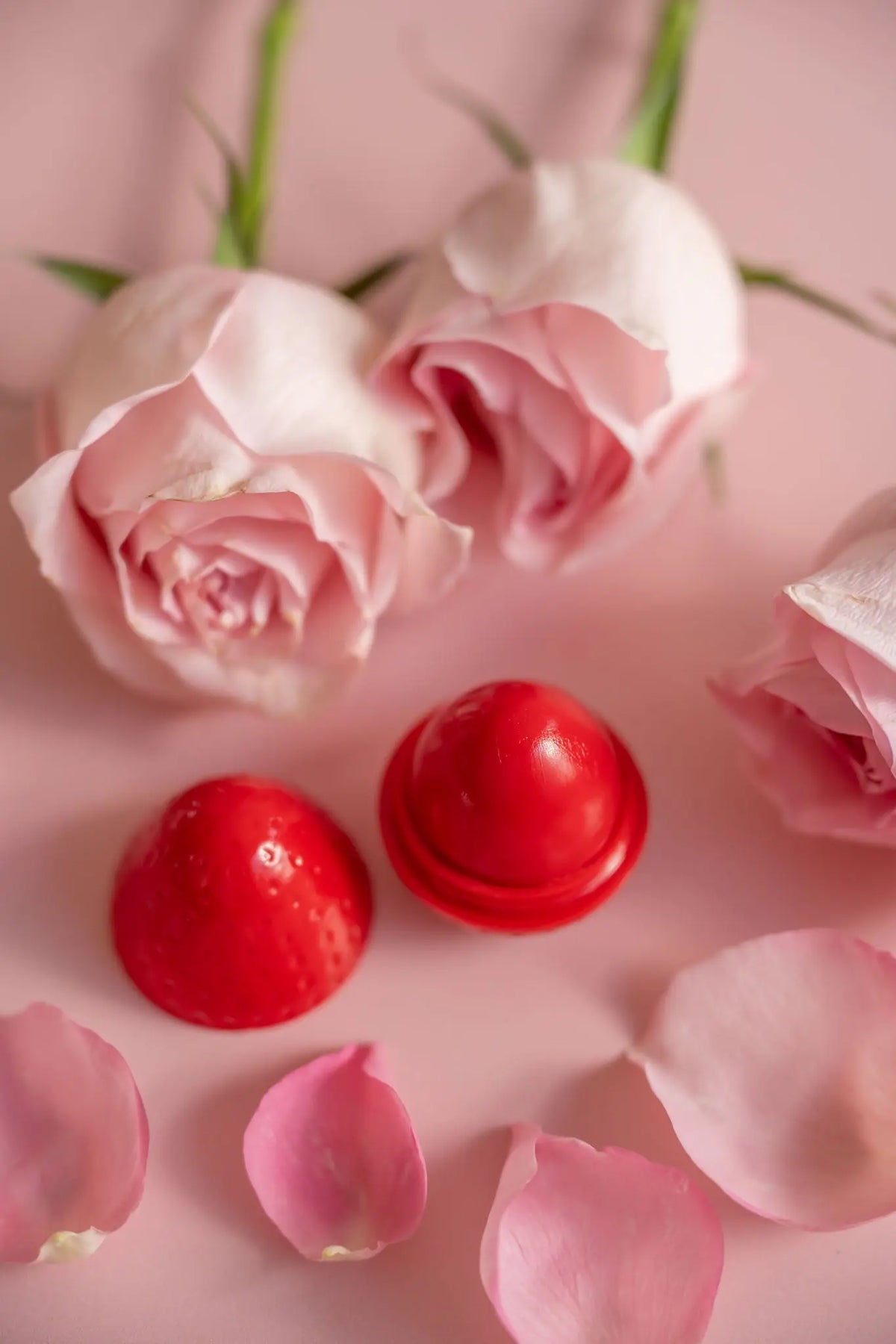 Red dome-shaped lip butter next to its lid, surrounded by soft pink roses and petals on a pastel pink background.