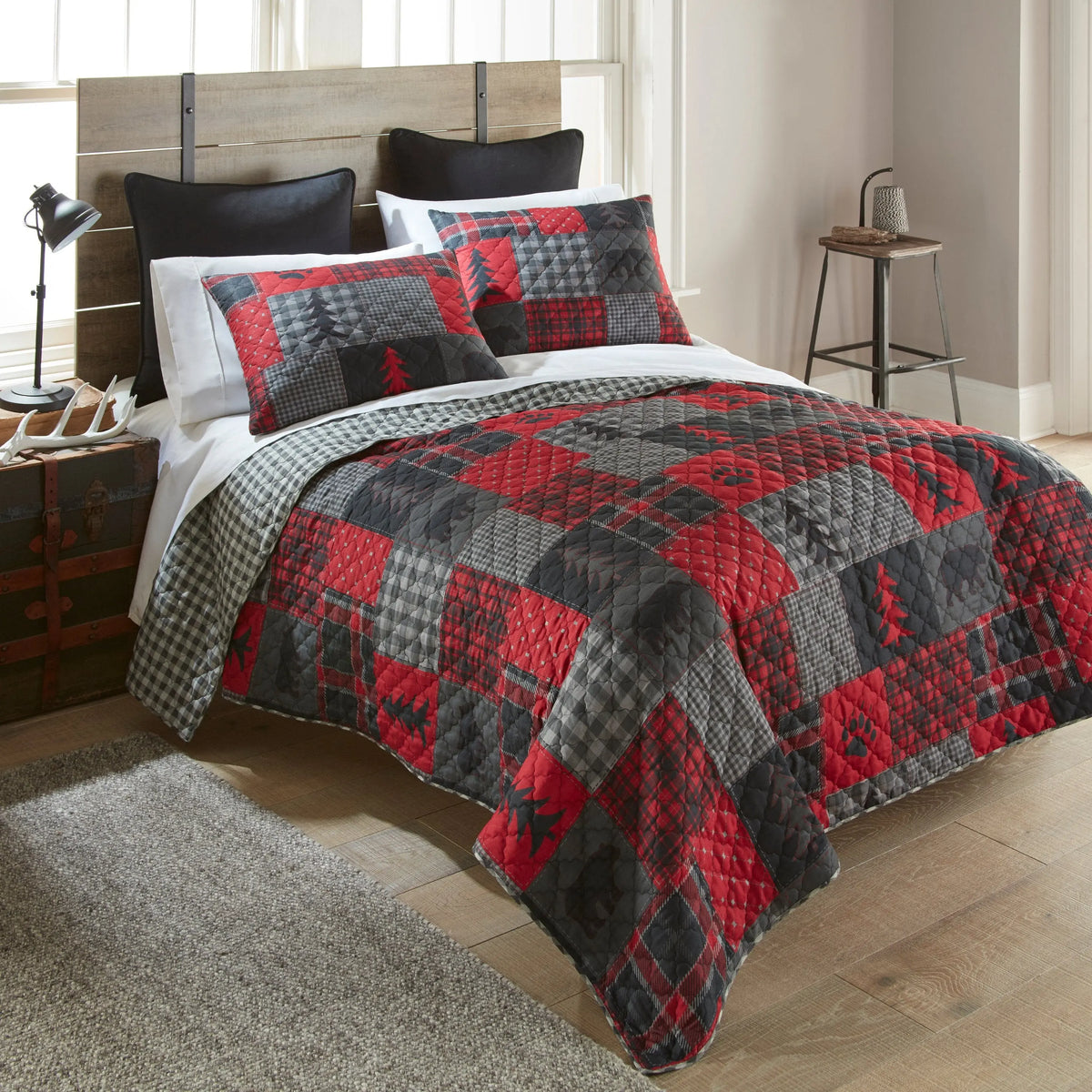 Rustic patchwork quilt set with red and grey plaid, bear, and pine tree patterns on a bed in a cabin-style room.