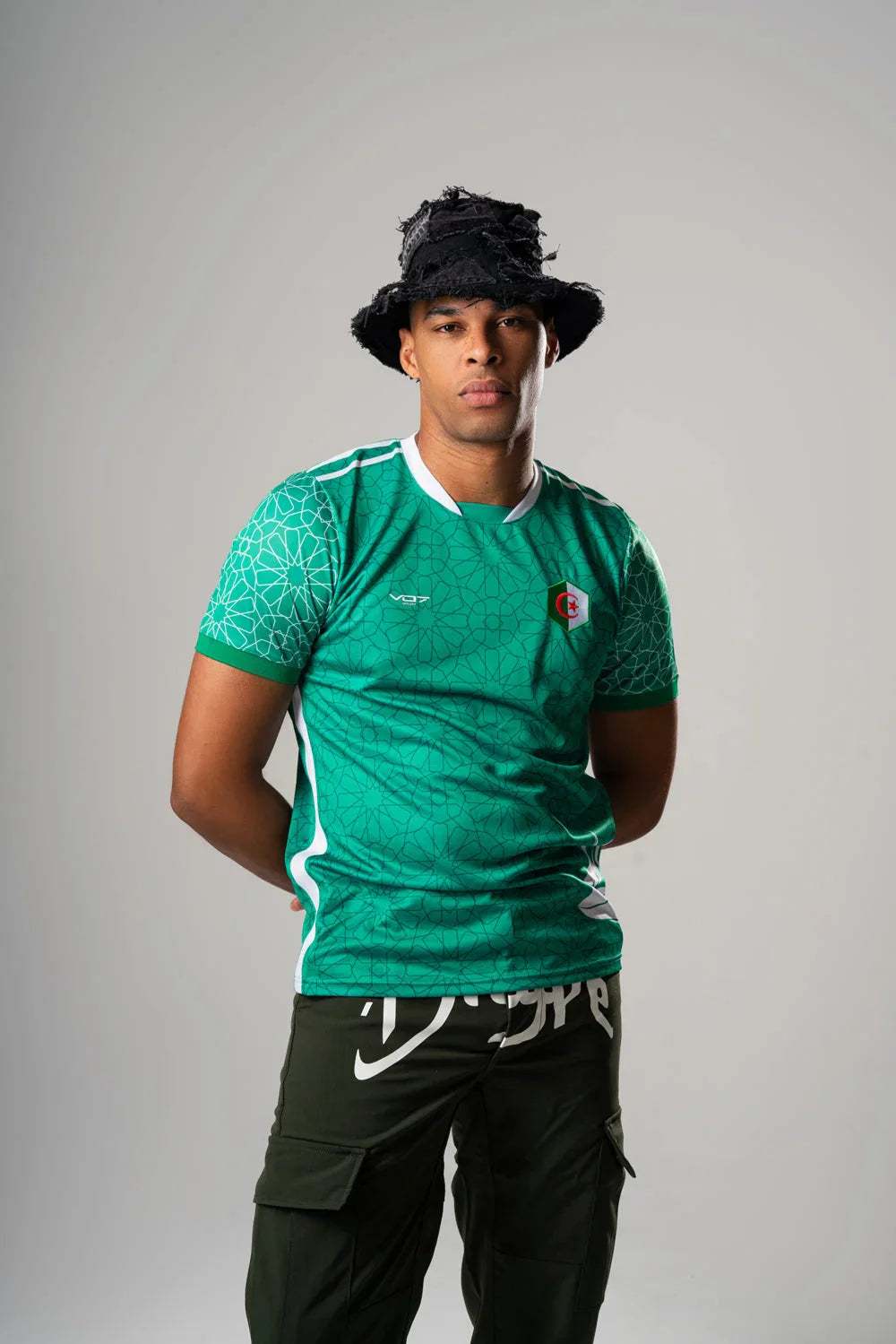 Mannequin portant un maillot VO7 vert à motifs géométriques, col blanc et écusson de l'Algérie sur fond gris.