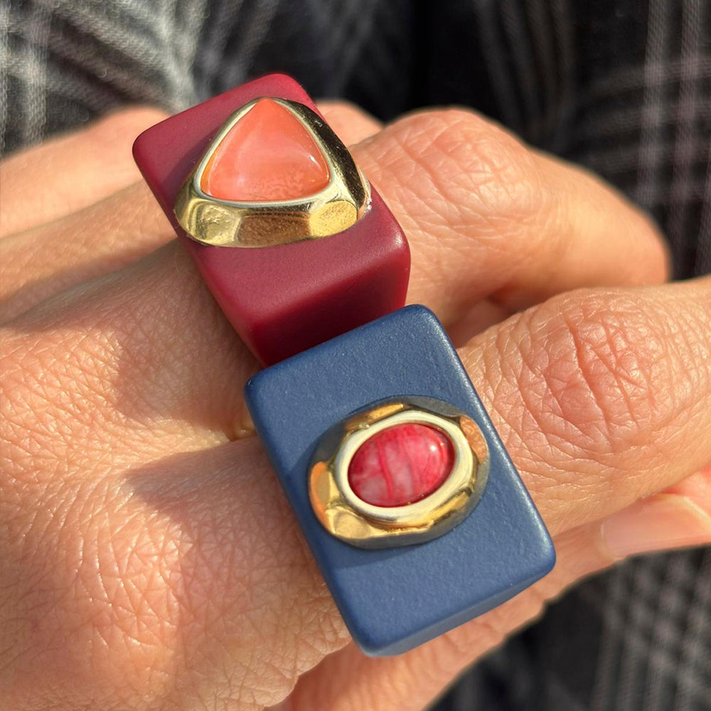 Anillo rectángulo azul con gema roja y anillo burdeos con piedra rosa, ambos con detalles dorados.