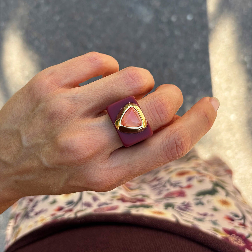 Anillo de resina burdeos con piedra triangular rosa y detalles dorados, estilo moderno y elegante.