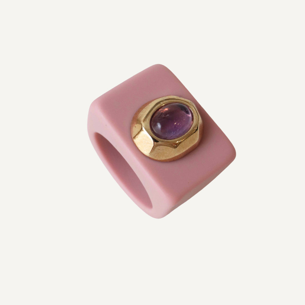 Anillo cuadrado de silicona rosa con gema morada y detalle dorado, estilo moderno y llamativo.