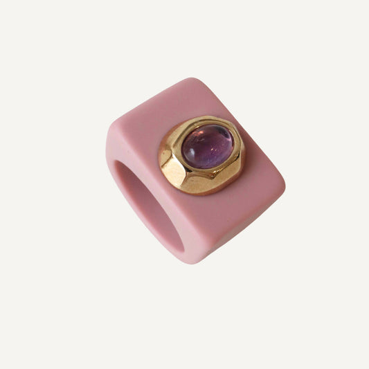 Anillo cuadrado de silicona rosa con gema morada y detalle dorado, estilo moderno y llamativo.