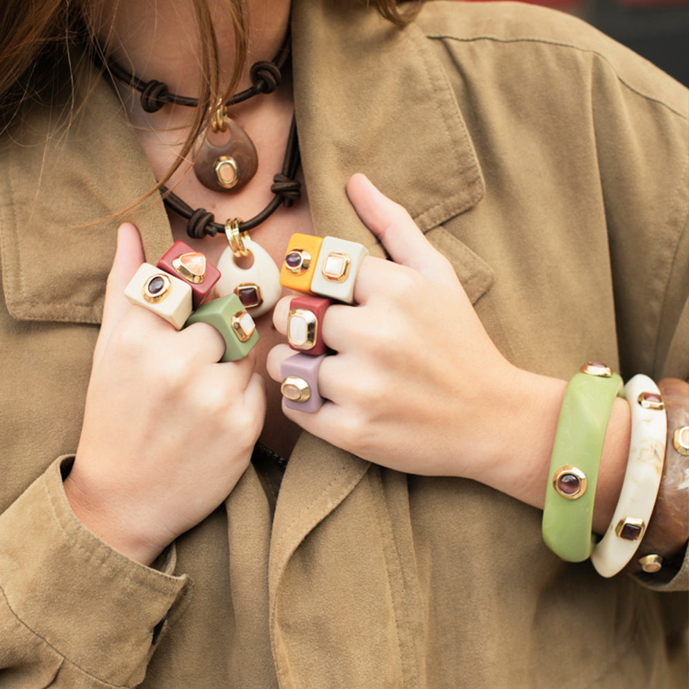 Anillos de resina en tonos verde, amarillo y púrpura, con detalles dorados, acompañados de pulseras de colores.