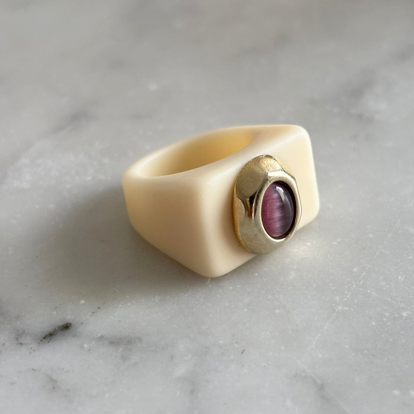 Anillo de silicona color crema con piedra púrpura y detalle dorado en forma de óvalo.