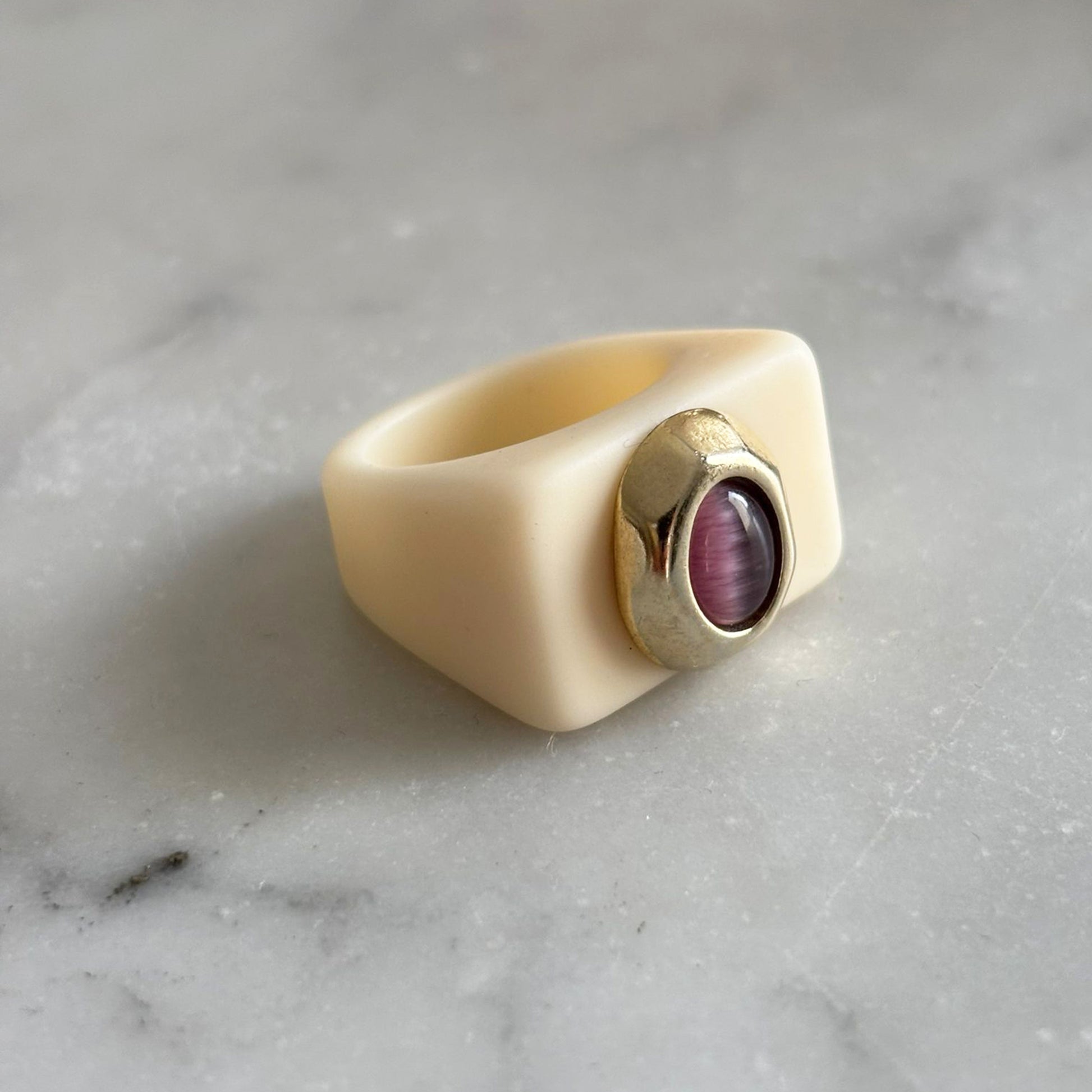 Anillo de silicona color crema con piedra púrpura y detalle dorado en forma de óvalo.
