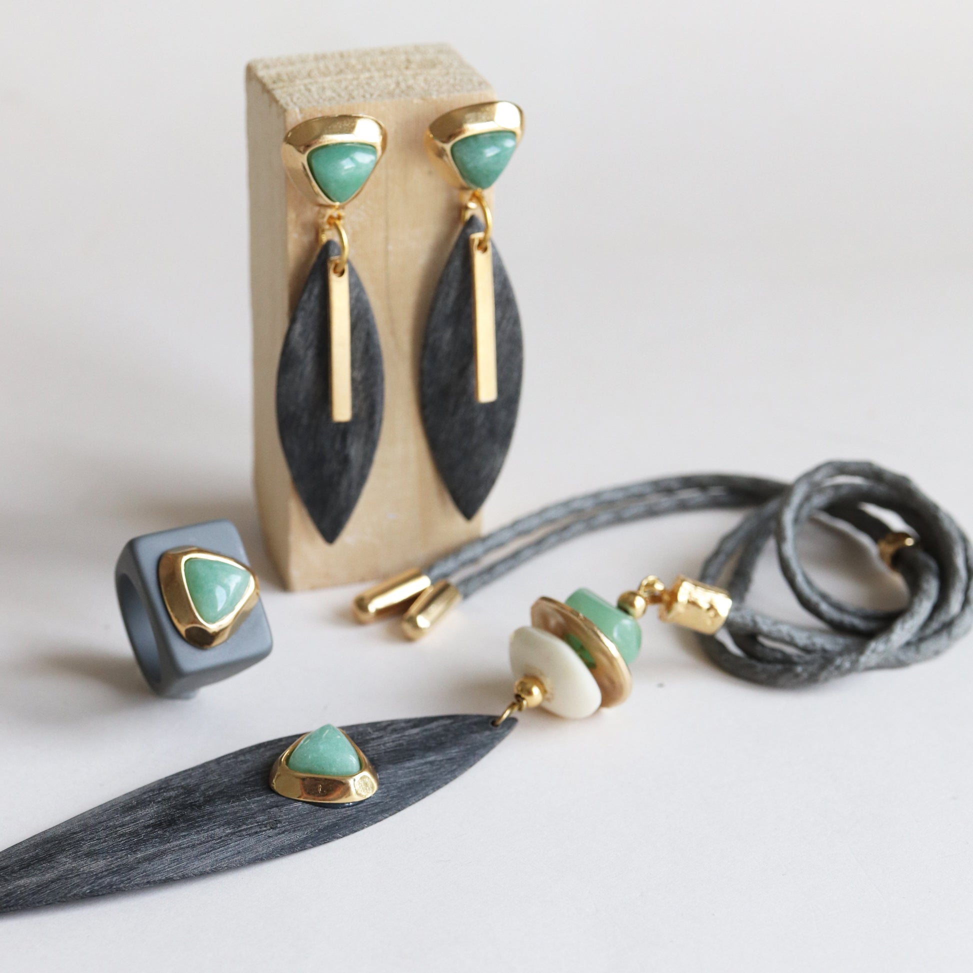 Pendientes dorados con piedra verde, anillo gris y collar con detalles en tonos verdes y madera, estilo moderno y elegante.