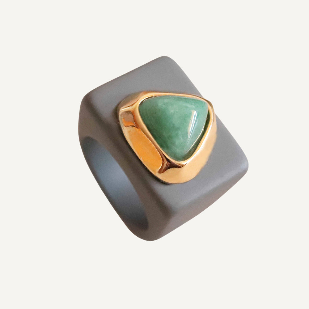 Anillo de cerámica gris con piedra verde triangular y detalle dorado. Estilo moderno y elegante.
