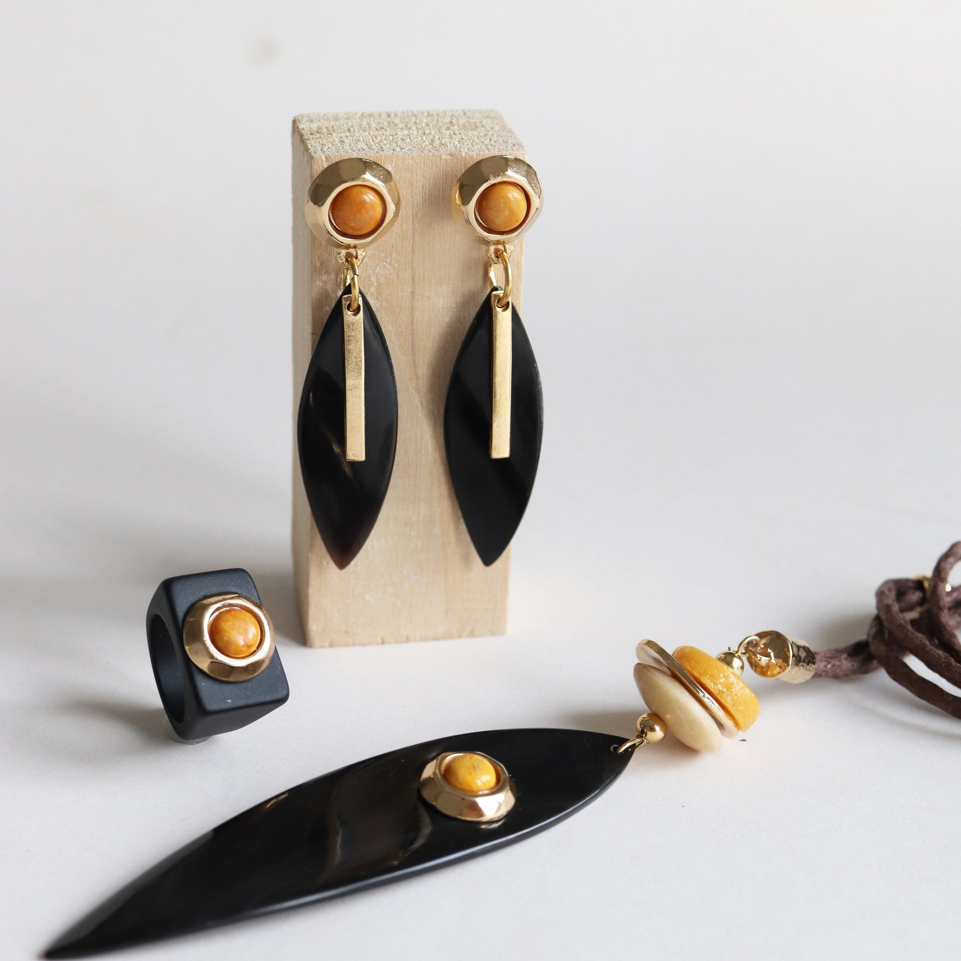 Pendientes negros con detalles dorados y anillo negro, collar con cuentas de madera y cuero, estilo moderno.