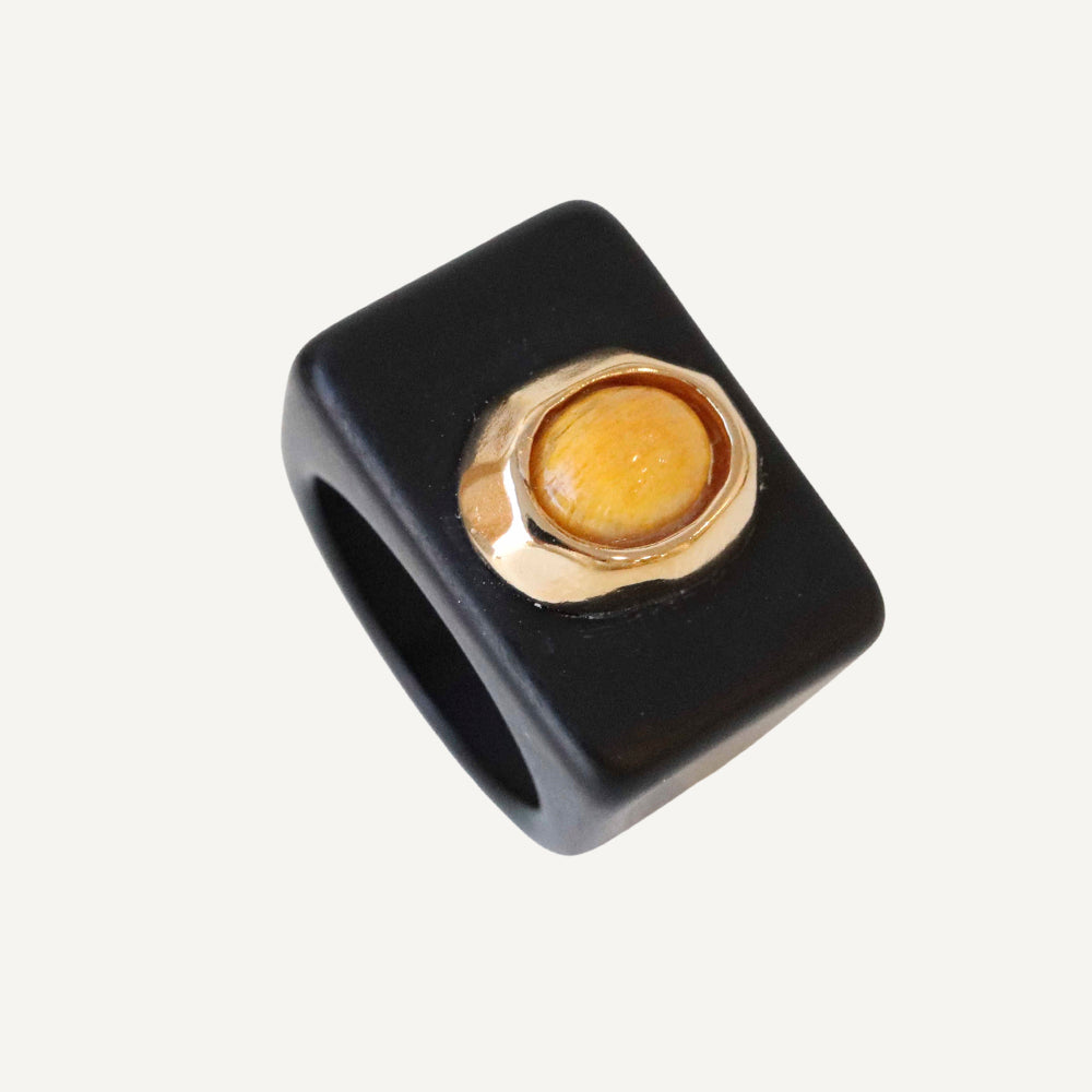 Anillo cuadrado negro de resina con detalle dorado y piedra amarilla en el centro. Estilo moderno y elegante.