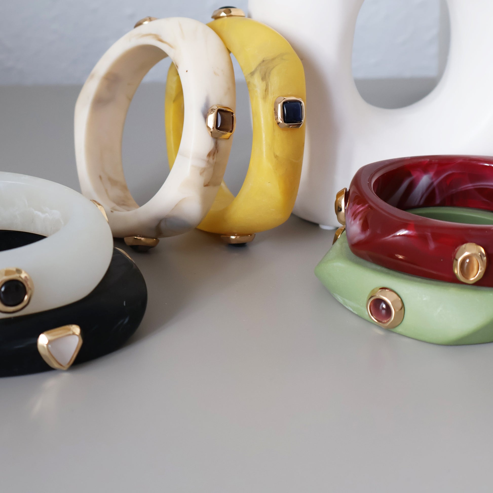 Pulseras de resina en colores blanco, amarillo, rojo, verde y negro, con detalles dorados en estilo moderno.