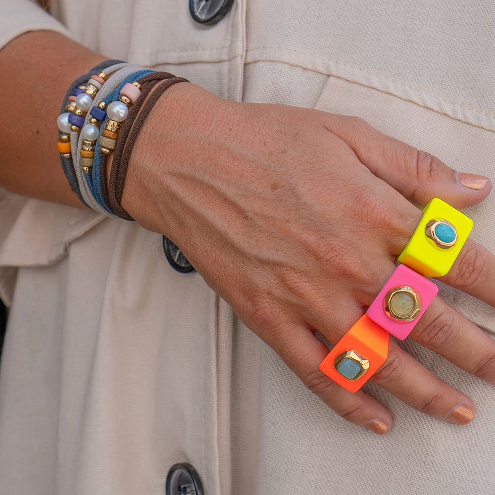 Anillo cuadrado rosa neón y amarillo con gema, pulseras de cuero y perlas en tonos marrones y azules.