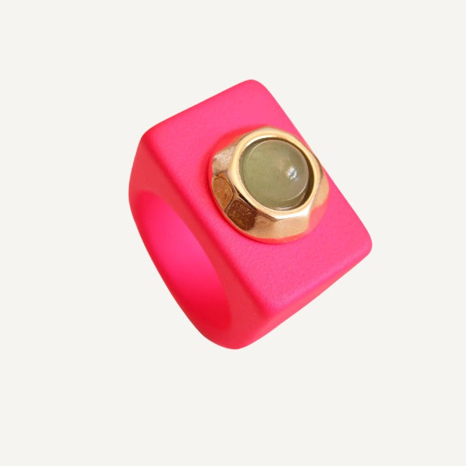 Anillo cuadrado de silicona rosa con detalle dorado y piedra verde. Estilo moderno y llamativo.