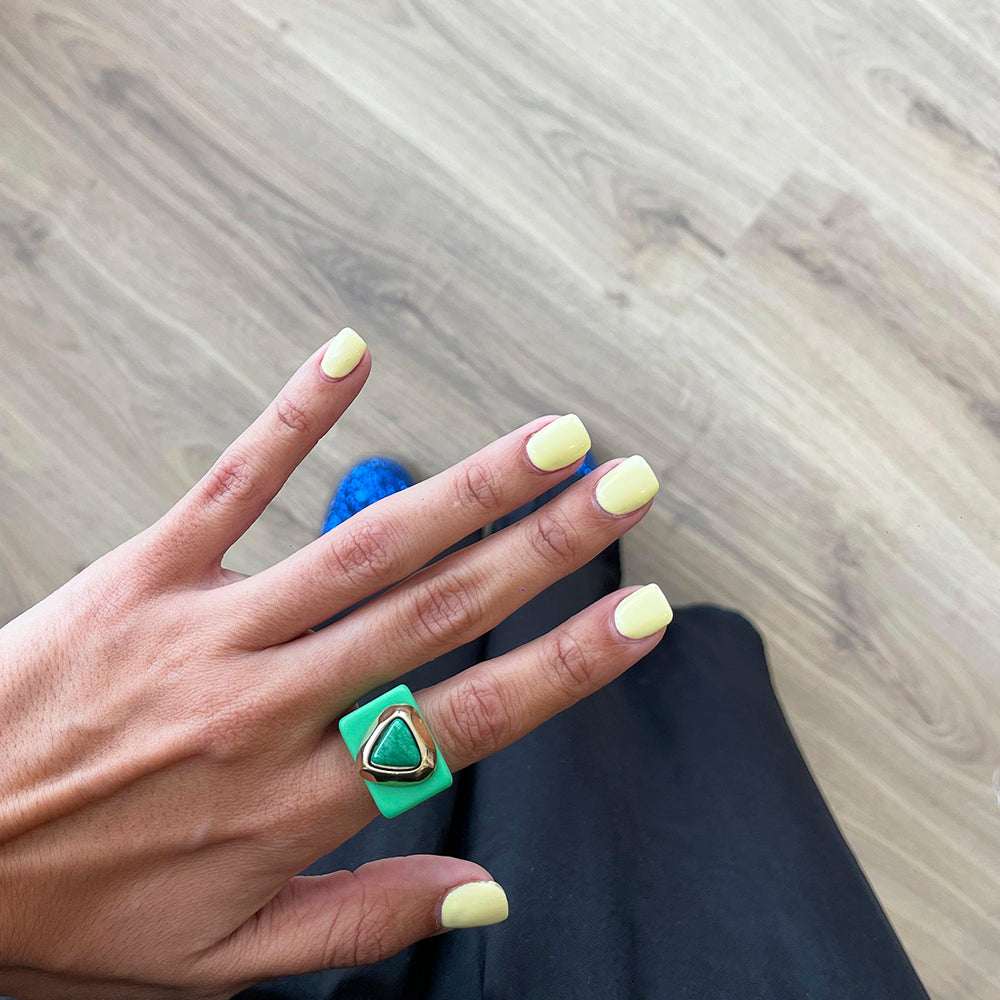 Anillo verde con forma triangular, piedra dorada, fondo de esmalte de uñas amarillo claro. Estilo moderno y vibrante.