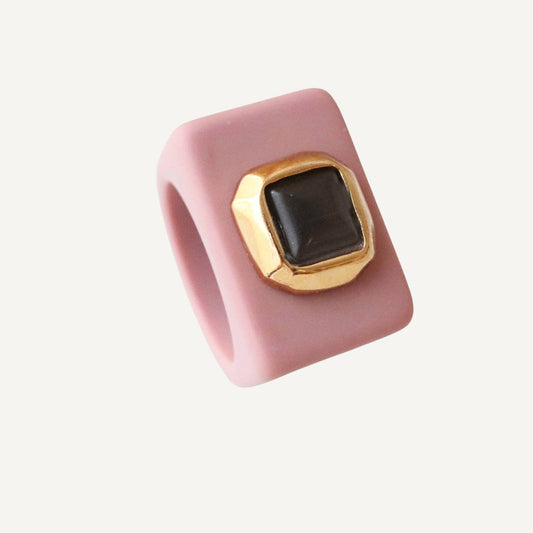 Anillo de silicona rosa con base dorada y piedra negra cuadrada. Diseño moderno y elegante.