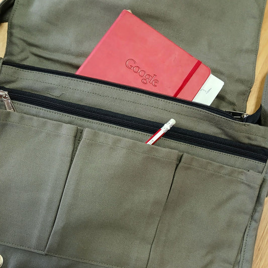 Bolsa mensajera color oliva de lona con bolsillos frontales, un cuaderno rojo y un lápiz en su interior.