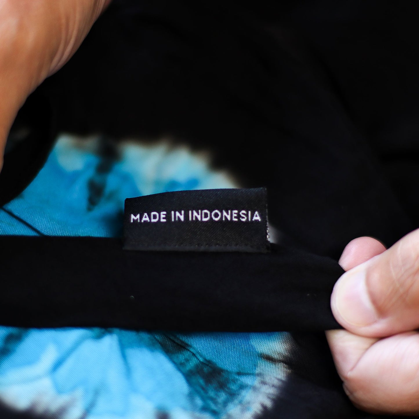 Mano sosteniendo la etiqueta negra con texto blanco "MADE IN INDONESIA" sobre tela azul y negra de estilo japonés.