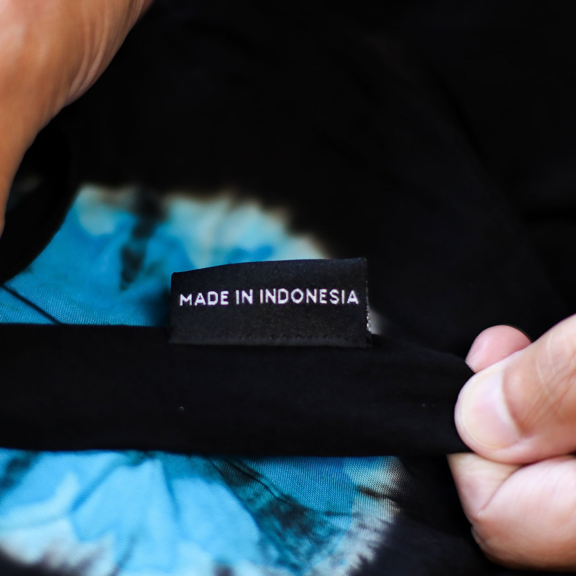 Mano sosteniendo la etiqueta negra con texto blanco "MADE IN INDONESIA" sobre tela azul y negra de estilo japonés.