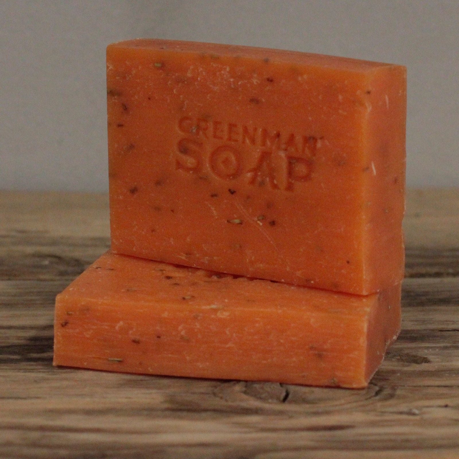 Dos pastillas de jabón naranja con motas, marcadas como "Greenman Soap", sobre una superficie de madera rústica.