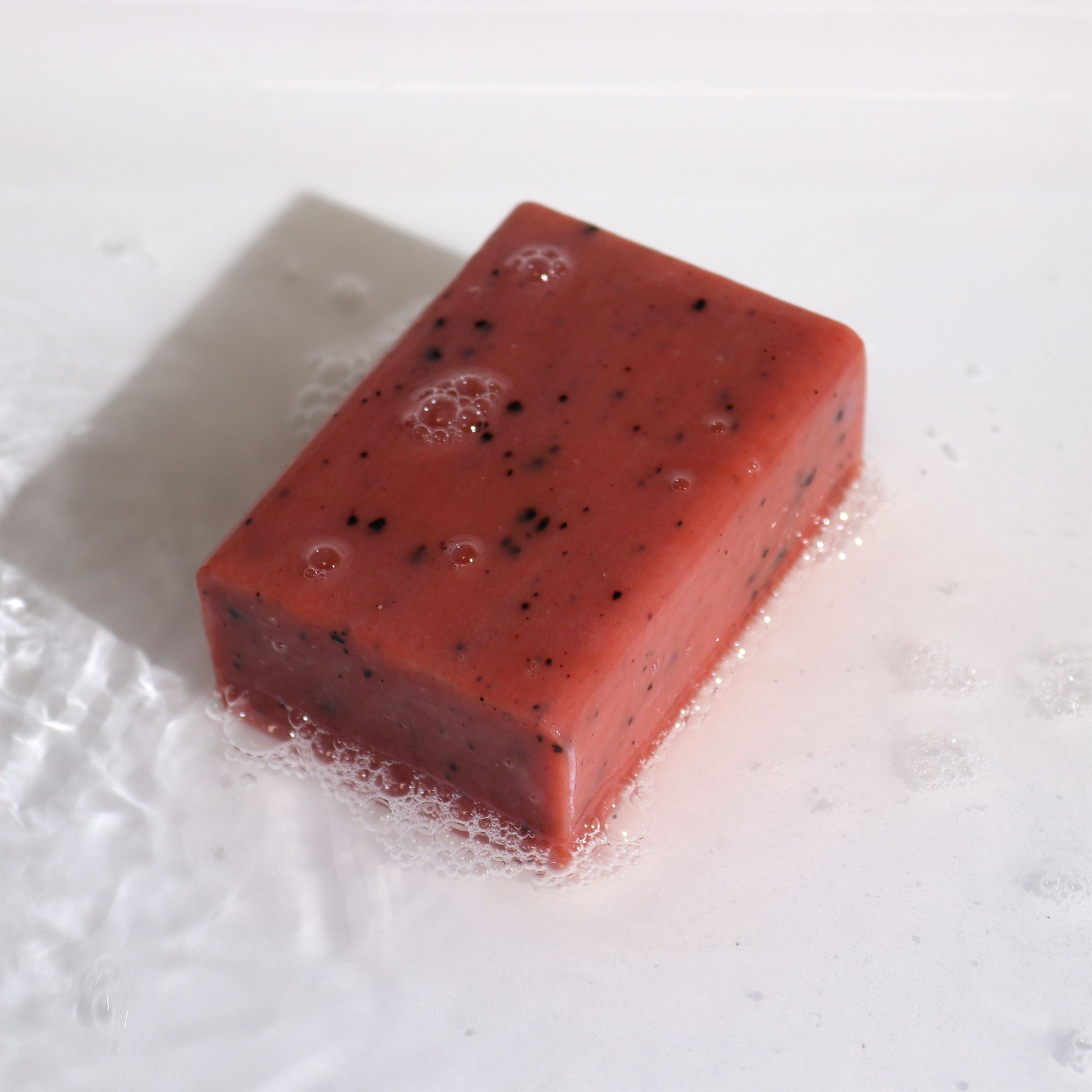 Jabón artesanal rosa con semillas de frutas, textura suave y burbujas, ideal para pieles sensibles.